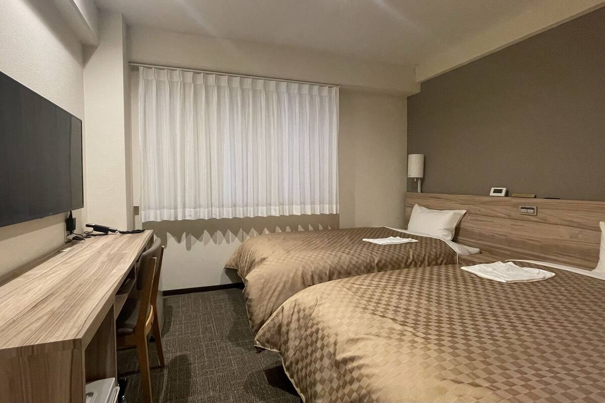 A hotel room with two beds, a desk, a television, and a window with curtains.