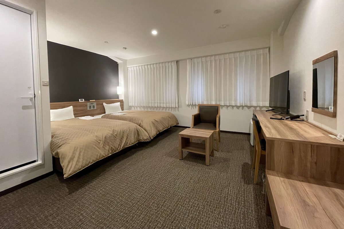 A hotel room with two beds, a desk, a chair, a TV, and a window with curtains.