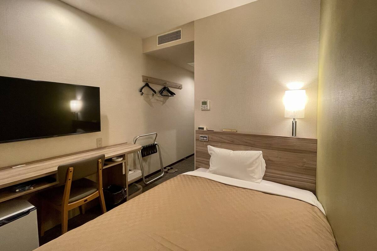 A hotel room with a bed, a desk with a chair, a television, a bedside table, and a lamp.