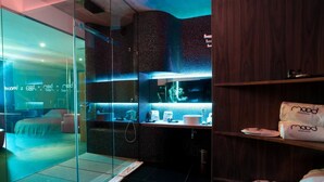 A modern bathroom with a glass shower enclosure, a wooden vanity, and a bed visible through the glass. - Mood - Private Suites (Montijo)