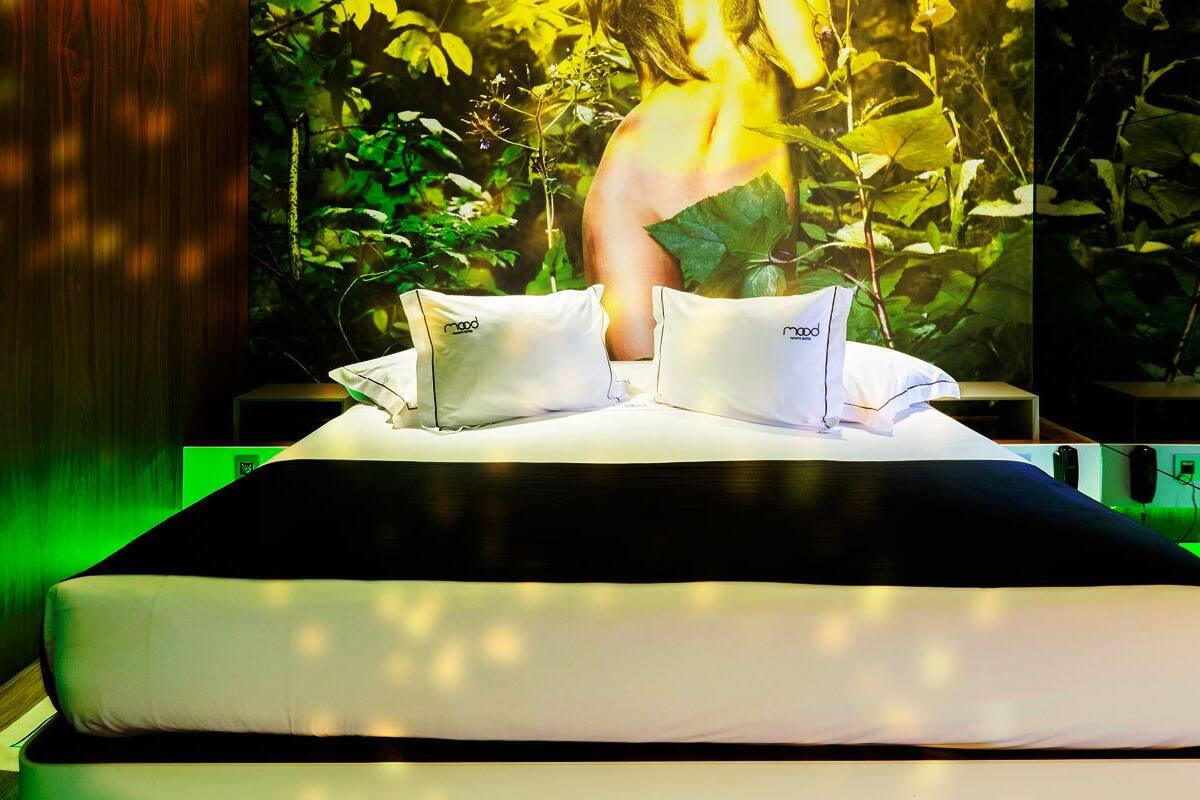 A bed with white bedding and two pillows, set against a backdrop of lush green foliage and a large plant.