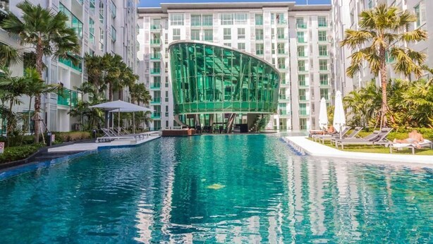 A hotel with a large swimming pool, palm trees, and a modern building with a curved glass facade. - City Center Residence by PSR ASIA (Pattaya)