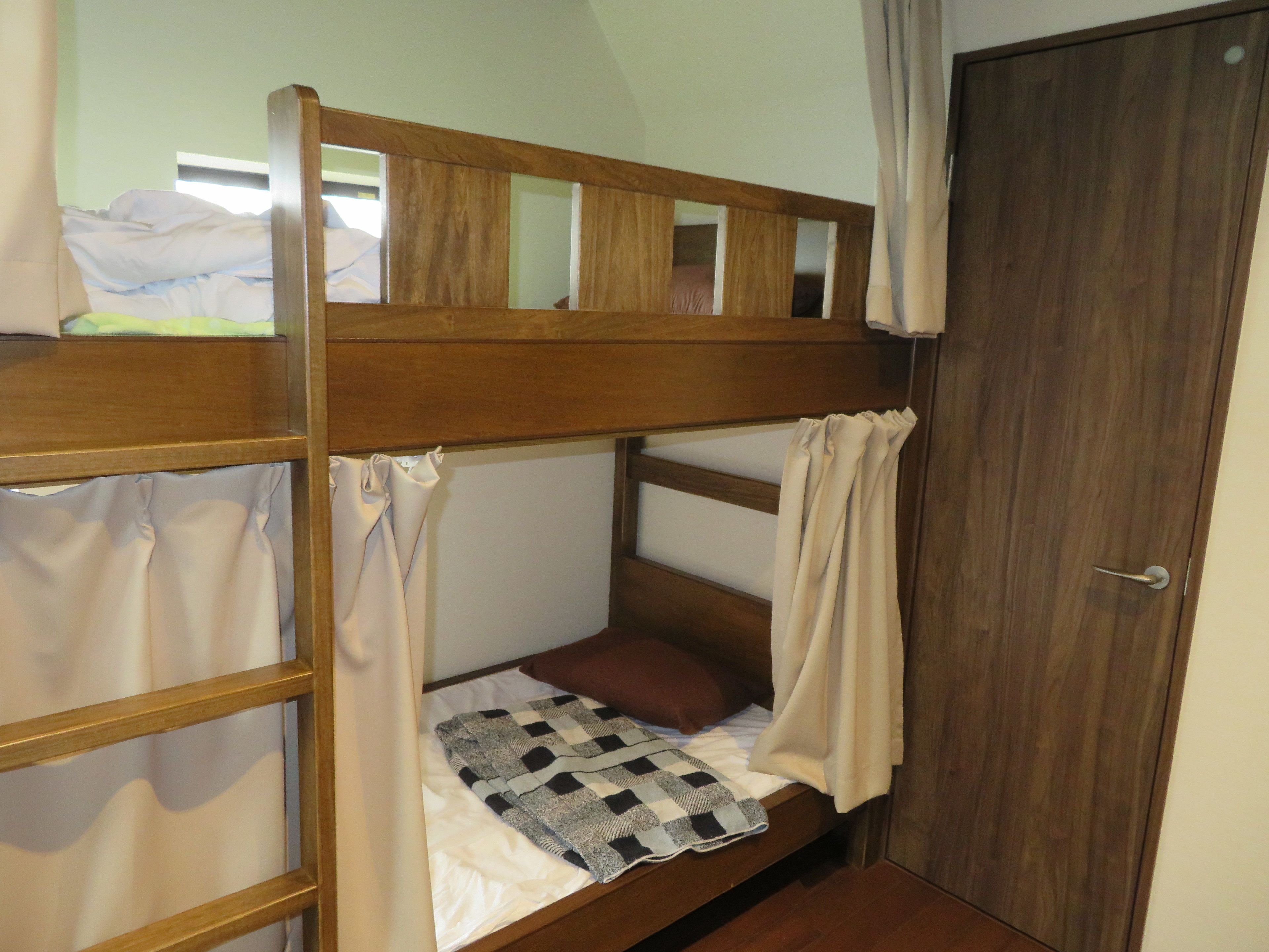 Un lit superposé avec une structure en bois, un matelas blanc et une couverture à carreaux. Il y a une porte sur la droite et une fenêtre au-dessus du lit.