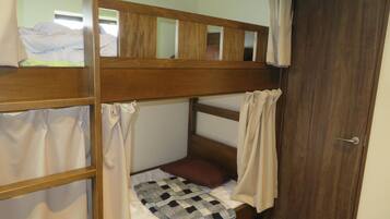 Un lit superposé avec une structure en bois, un matelas blanc et une couverture à carreaux. Il y a une porte sur la droite et une fenêtre au-dessus du lit.