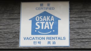 A sign for Osaka Stay Vacation Rentals with a blue house logo and text in English and another language. - Azalea - Hostel (Osaka)