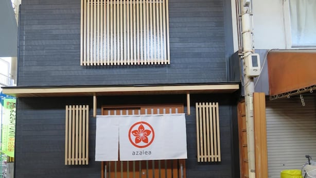 A building with a dark facade, a wooden gate, and a sign with a red flower logo. - Azalea - Hostel (Osaka)