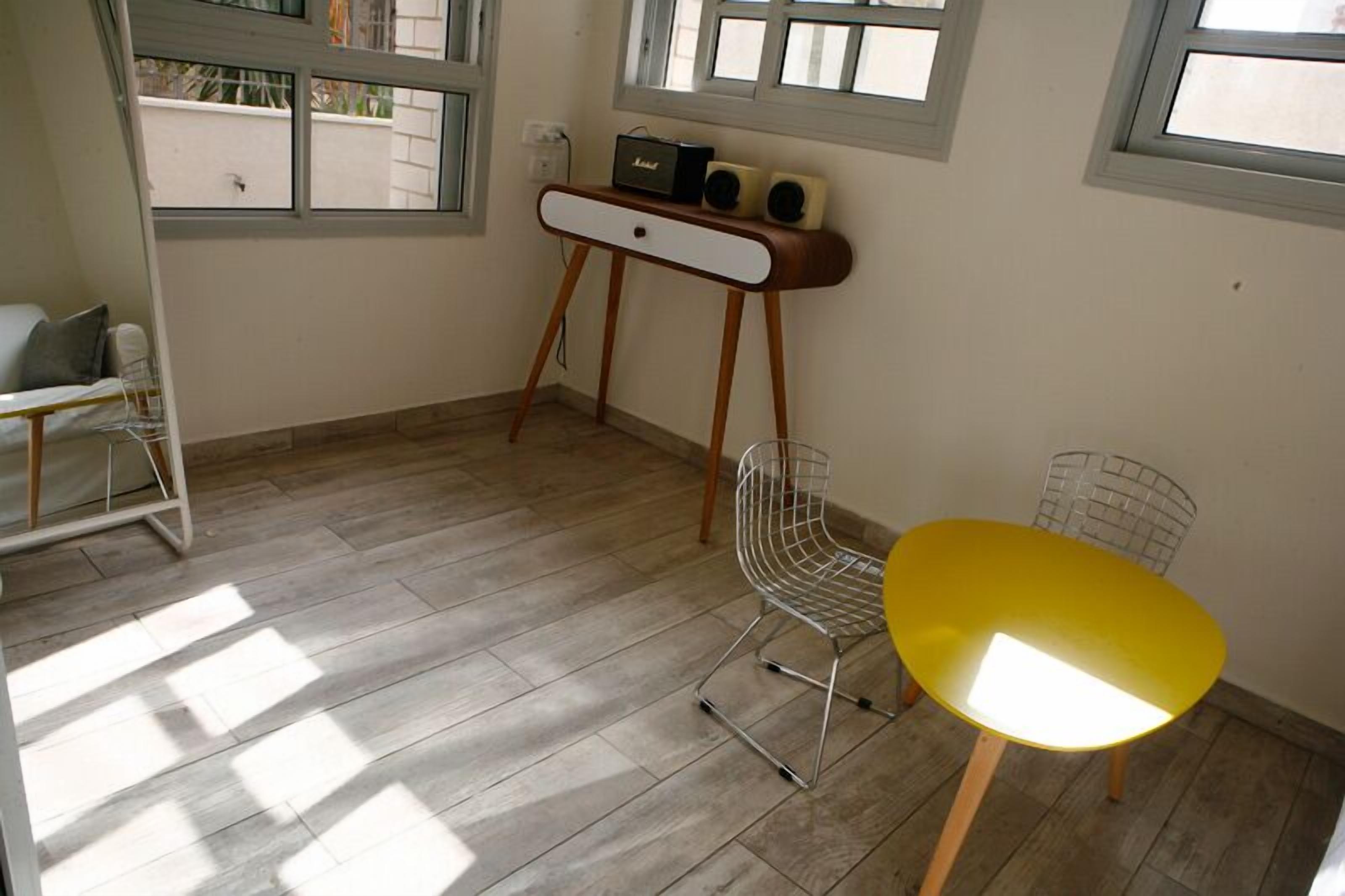A small room with a wooden floor, a yellow table, a white chair, and a small white desk with speakers.