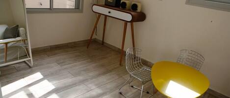 A small room with a wooden floor, a yellow table, a white chair, and a small white desk with speakers.