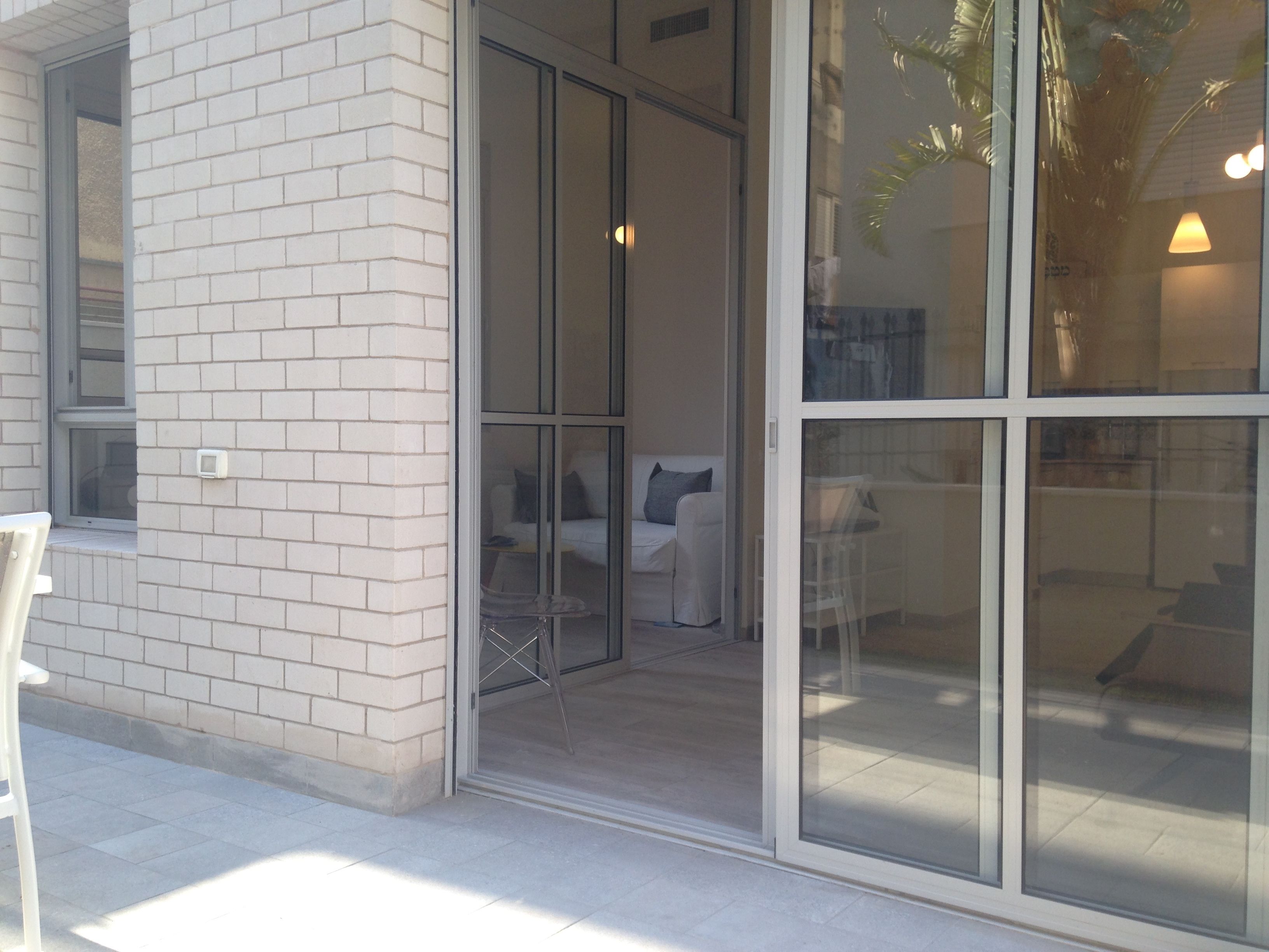 A modern building with a glass door entrance, a white brick wall, and a patio area with a white chair and a table.