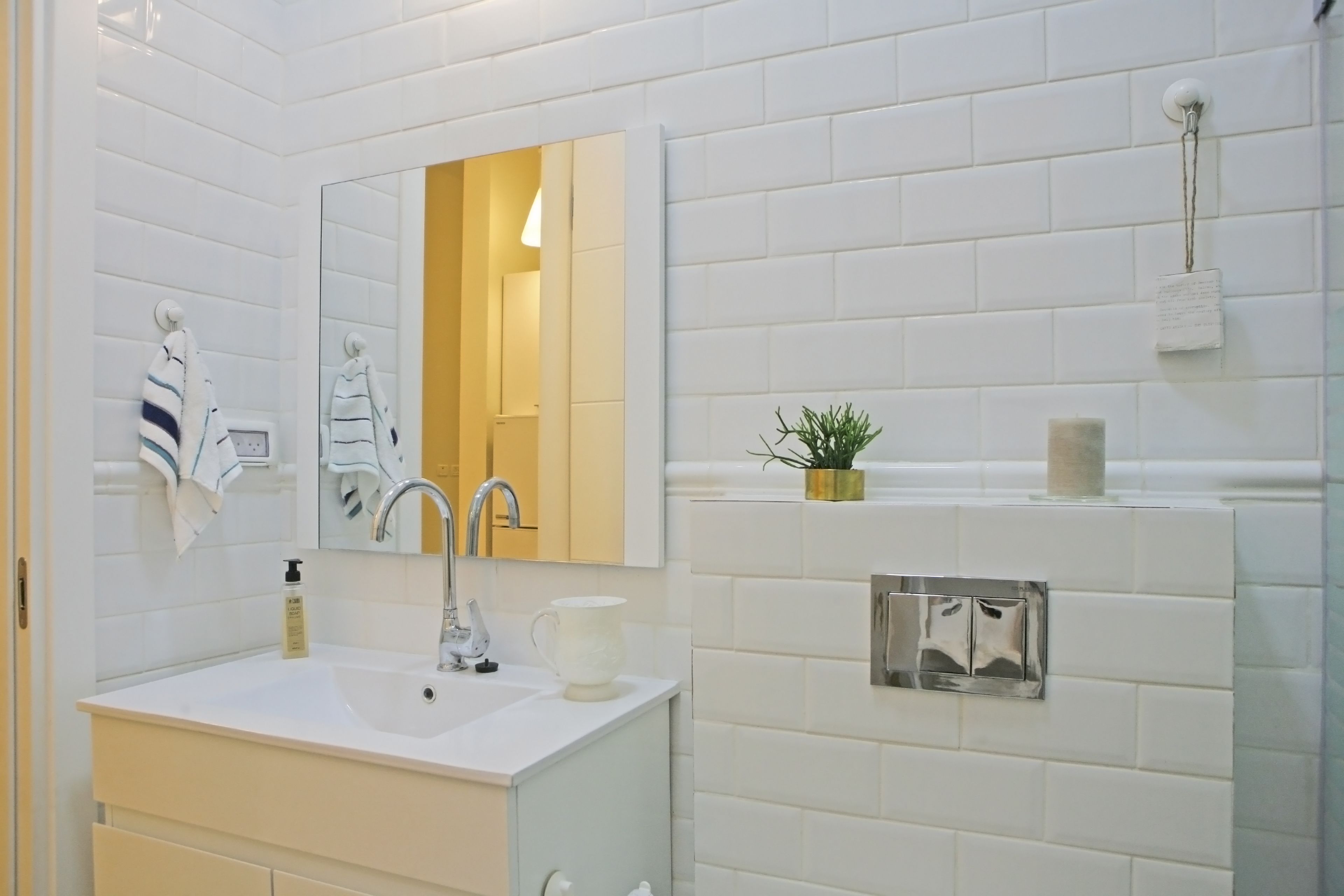 Een moderne badkamer met witte betegelde wanden, een grote spiegel, een wastafel met kraan, een handdoekrek, een kleine plant en een toiletrolhouder.