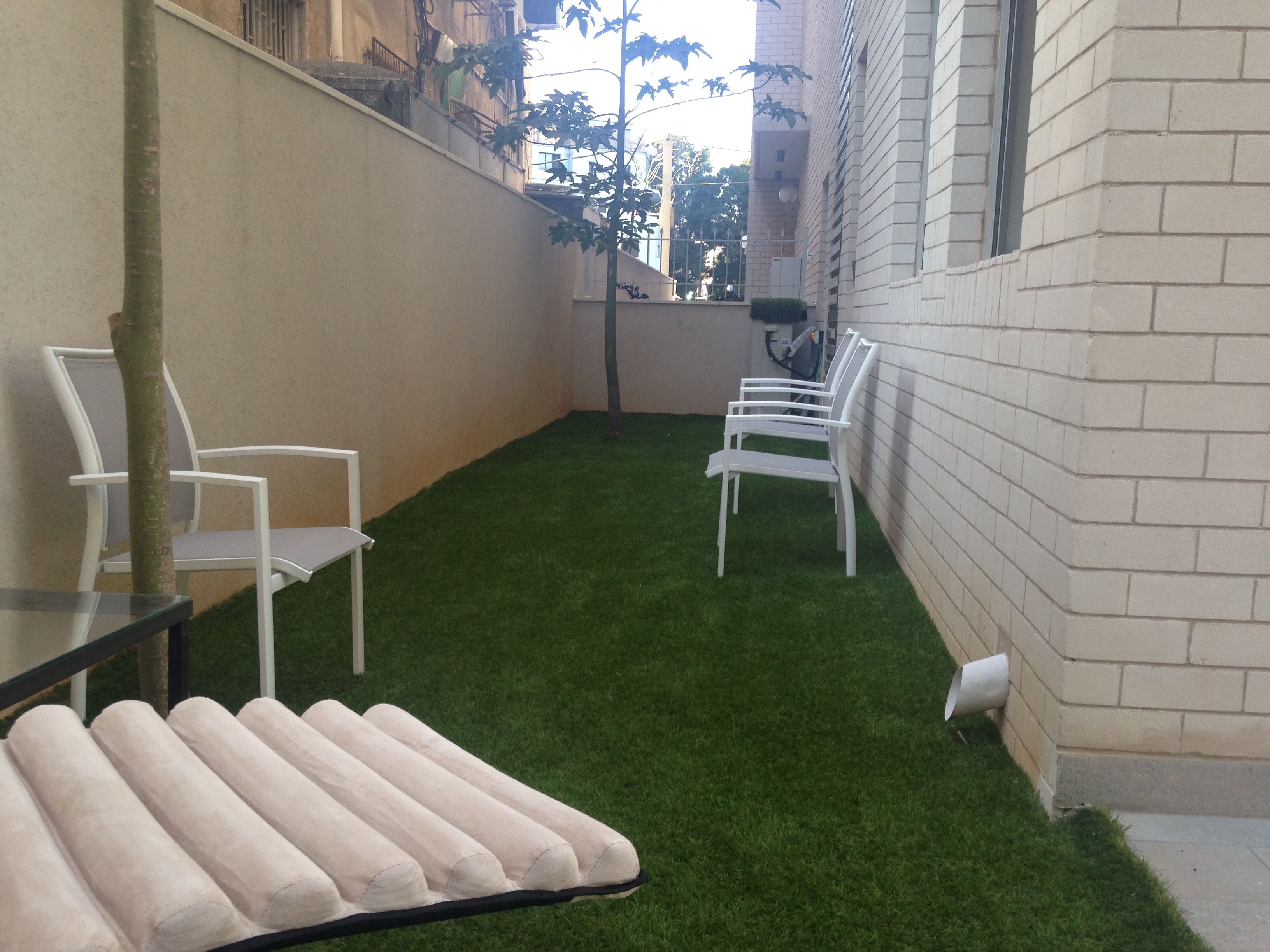 Un petit espace extérieur clos, doté d’une pelouse artificielle, de chaises blanches et d’une table sur laquelle sont posés des tapis enroulés.