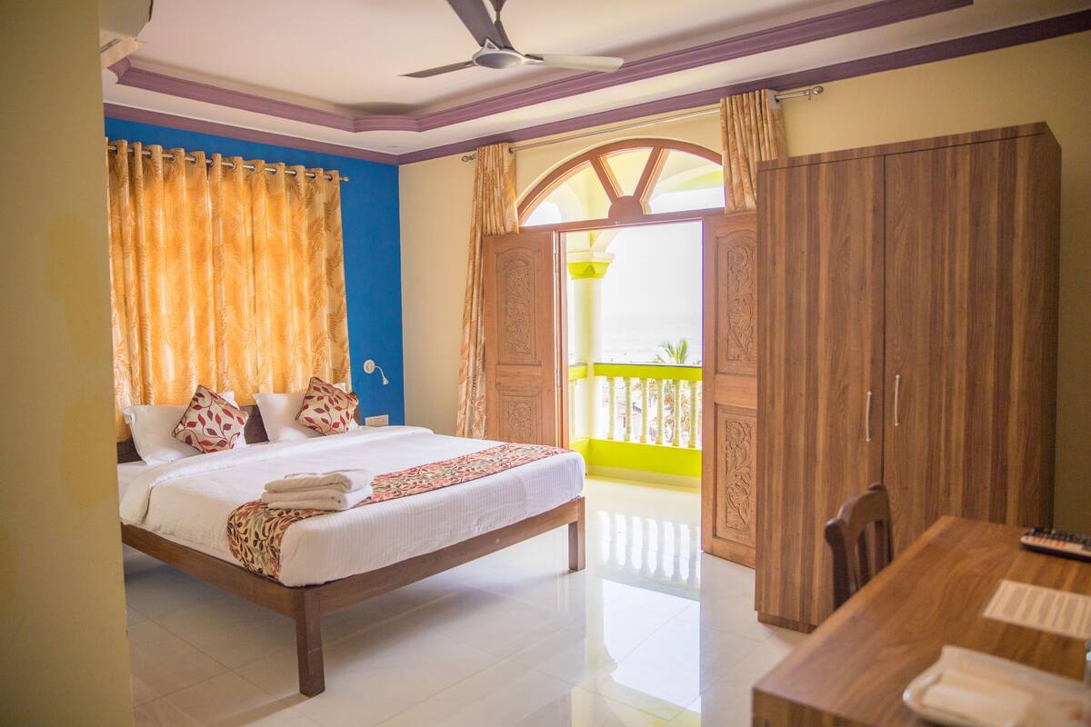 A bedroom with a bed, a wooden wardrobe, a dining table, and a balcony with a yellow railing.