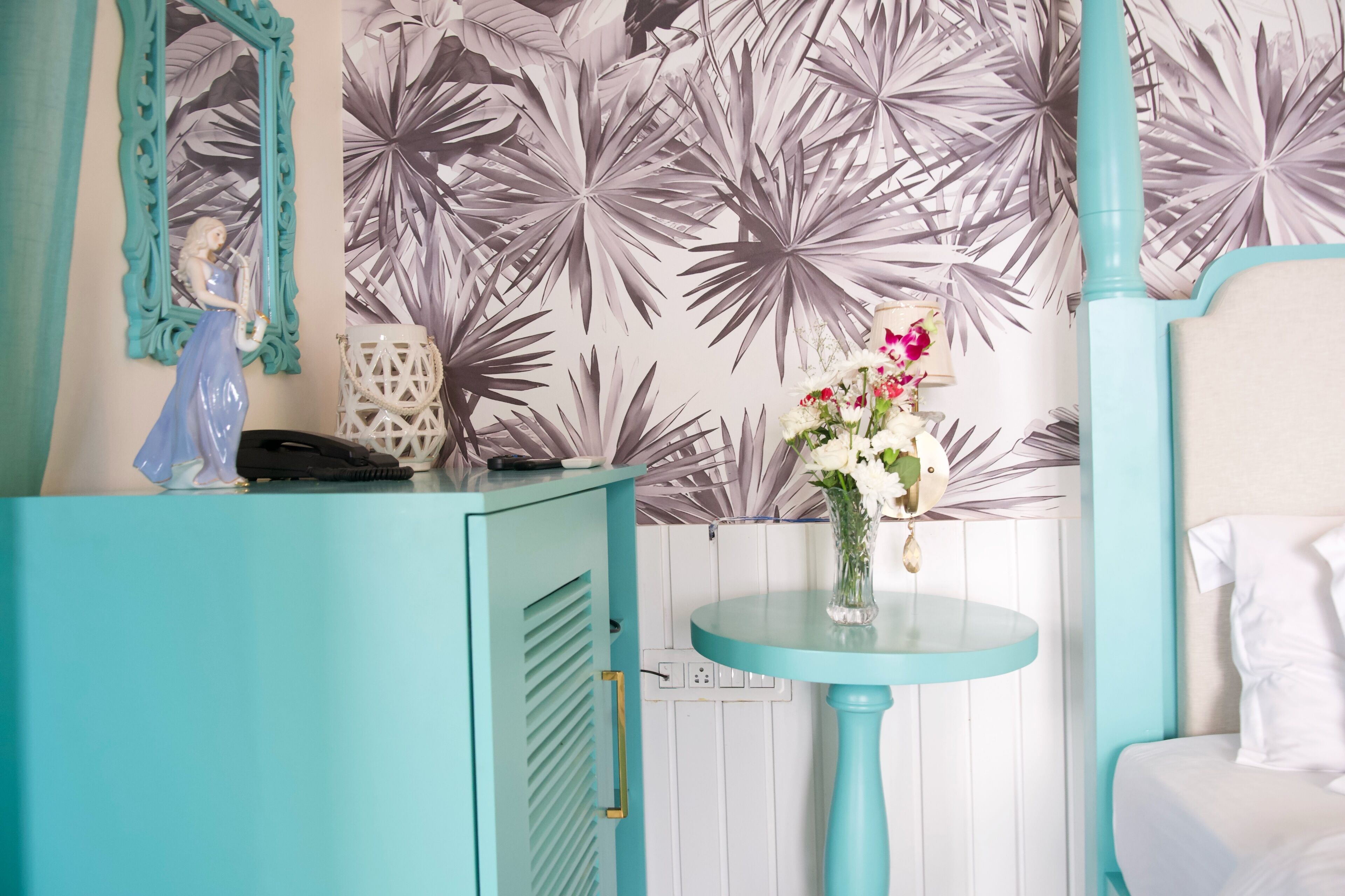 Una habitación con un armario azul verdoso, una mesa redonda con un florero y una pared con estampado de hojas tropicales.