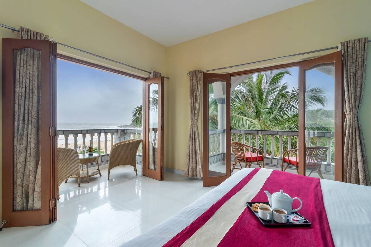 A hotel room with a bed, a balcony with wicker chairs and a table, and a view of palm trees and the sea.