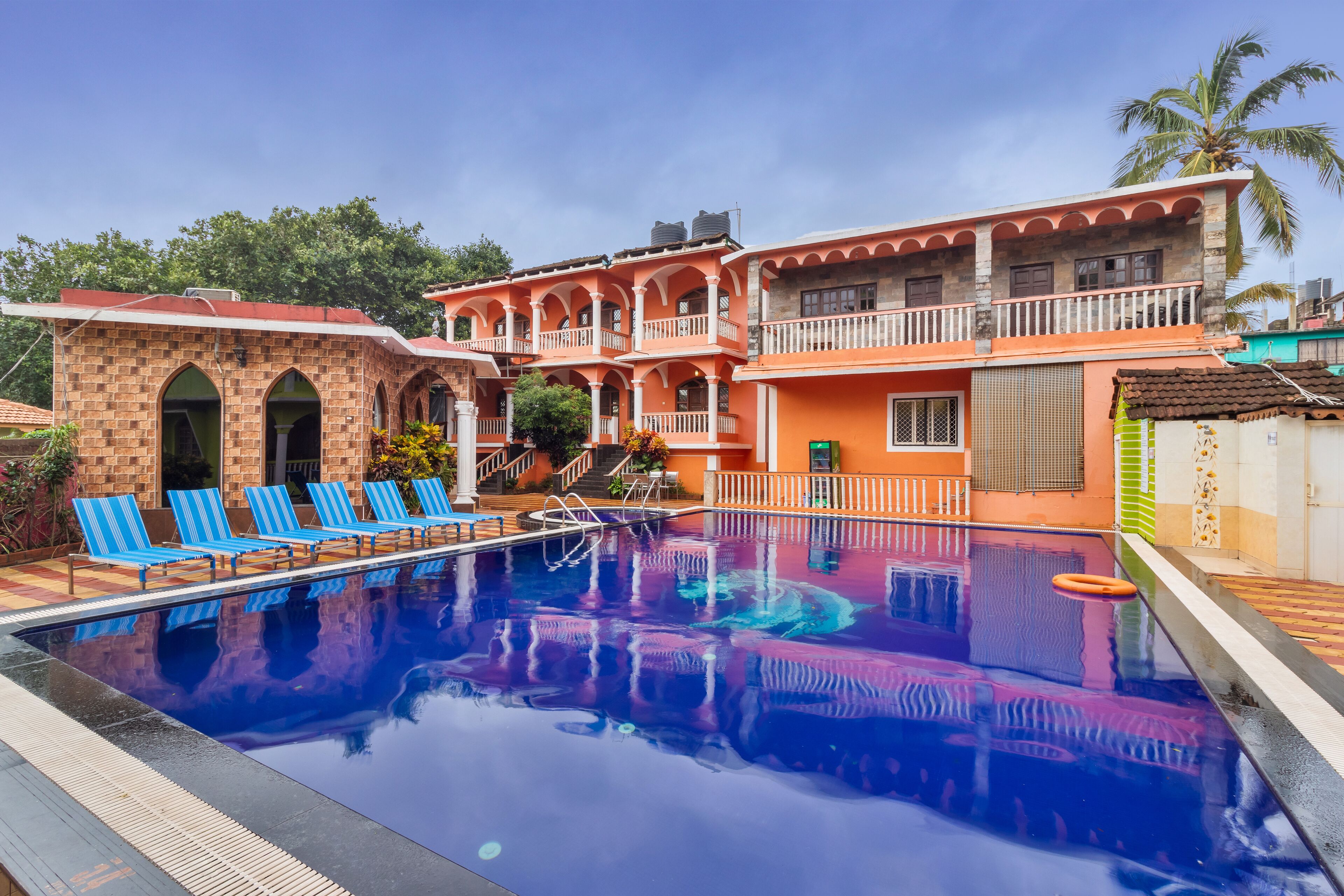 A swimming pool with blue lounge chairs, a building with arches and balconies, and a palm tree in the background.