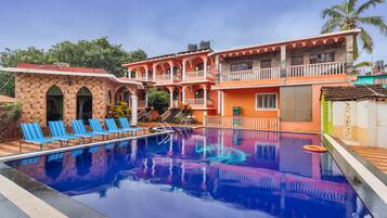 A swimming pool with blue lounge chairs, a building with arches and balconies, and a palm tree in the background.