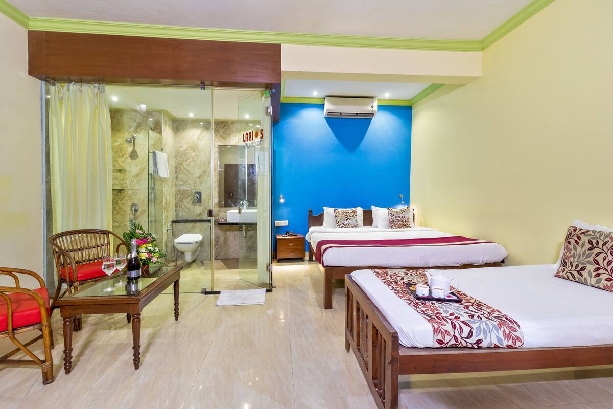 A hotel room with a glass shower enclosure, a wooden bed with a red and white bedspread, a wooden chair with a red cushion, and a small table with a bottle of champagne and glasses.