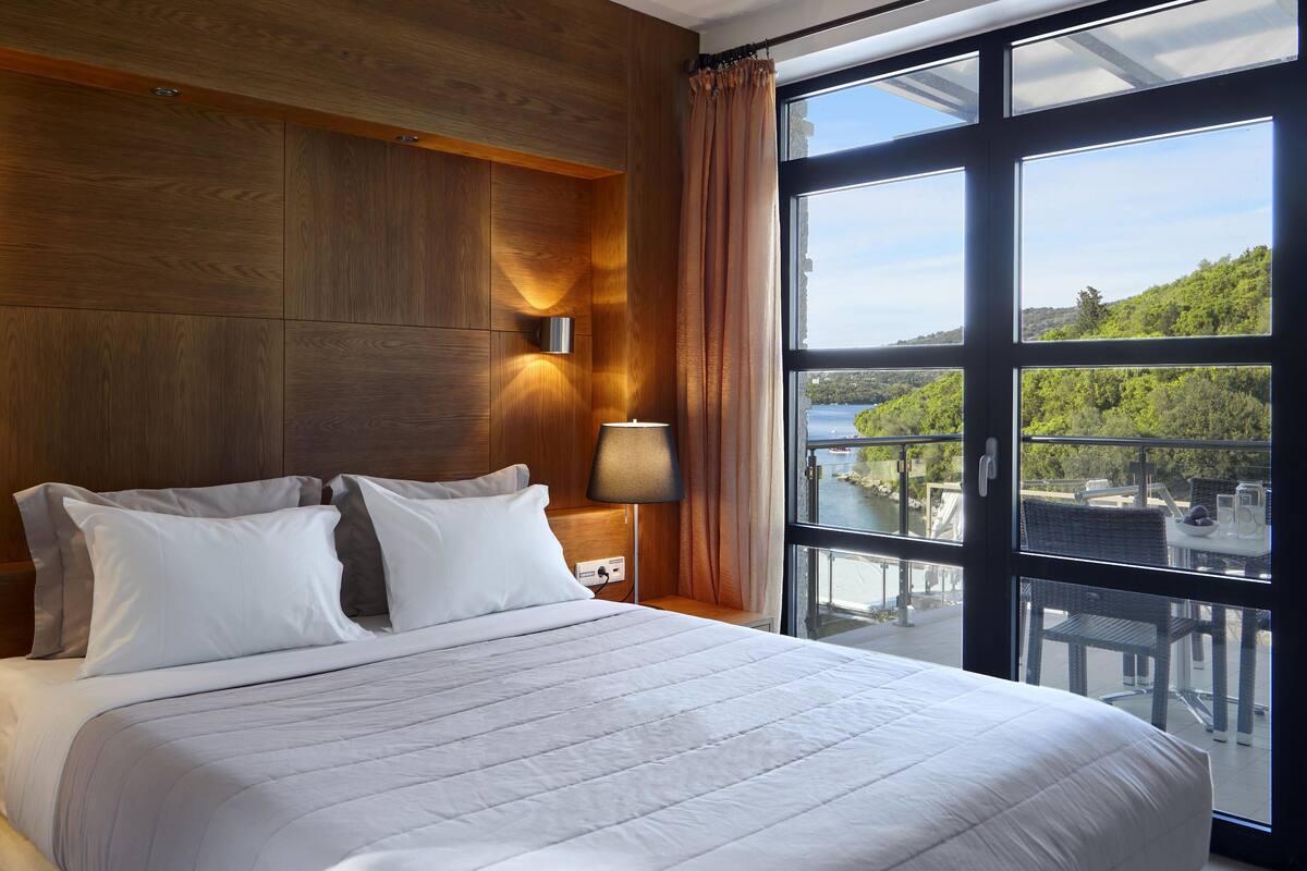 A hotel room with a large bed, wooden paneling, a nightstand with a lamp, and a view of a lake through large windows.