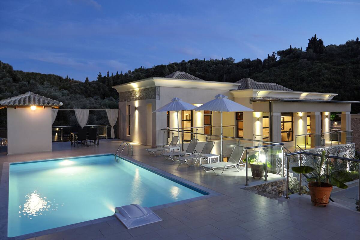 A swimming pool with lounge chairs and umbrellas, surrounded by a building with a tiled roof and a view of a forested hillside.