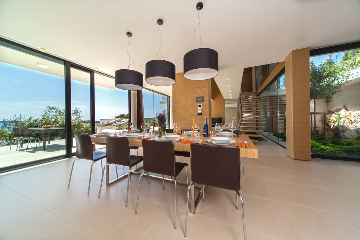 A modern dining area with a large table set for six, surrounded by chairs, and large windows offering a view of the outdoors.