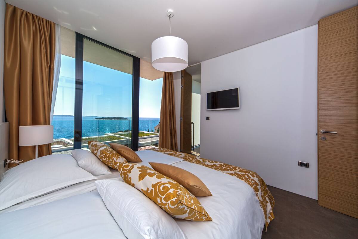 A modern hotel room with a large bed, a TV mounted on the wall, and a view of the ocean through floor-to-ceiling windows.