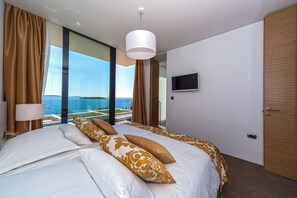A modern hotel room with a large bed, a TV mounted on the wall, and a view of the ocean through floor-to-ceiling windows. - Golden Rays Luxury Villas (Primosten)