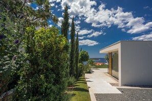 A modern building with a concrete pathway leading to a glass door, surrounded by lush greenery and tall trees. - Golden Rays Luxury Villas (Primosten)