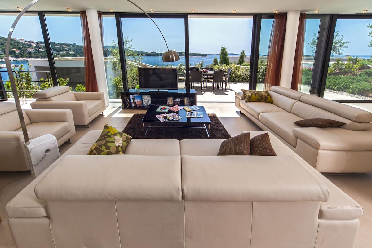 A modern living room with a large sofa, a coffee table, and a TV. There is a view of the sea through large windows.