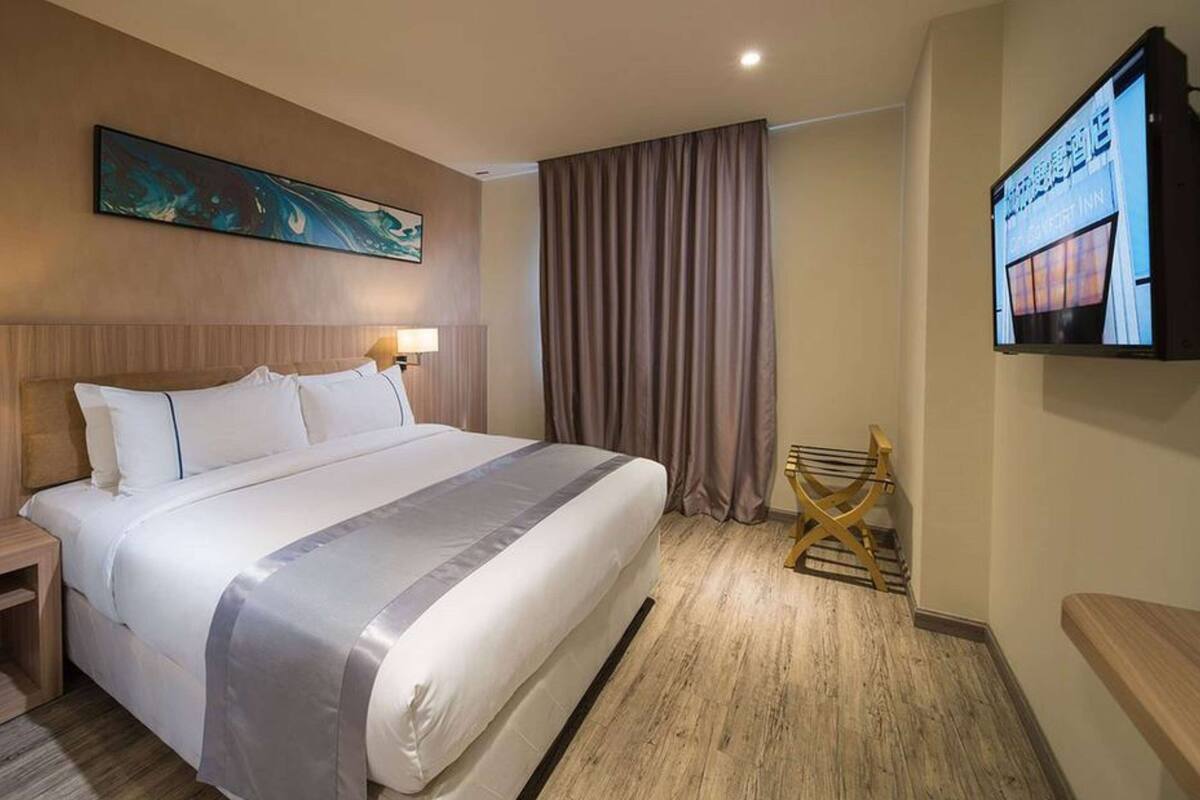 A hotel room with a large bed, a flat-screen TV mounted on the wall, a wooden floor, and a painting above the bed.