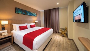Une chambre d’hôtel avec un grand lit, un téléviseur fixé au mur, une table de chevet en bois et une couvre-lit rouge et blanche.
