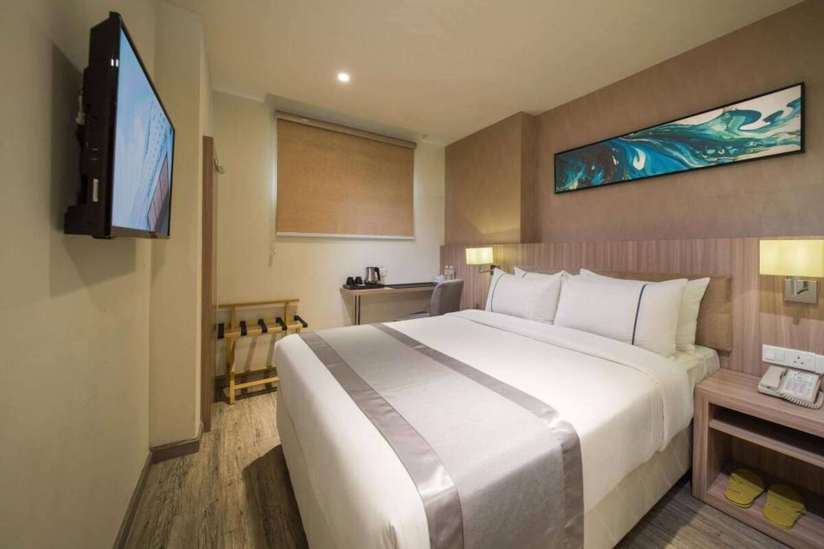 A hotel room with a large bed, a flat-screen TV mounted on the wall, a desk with a chair, a painting on the wall, and a telephone on the nightstand.