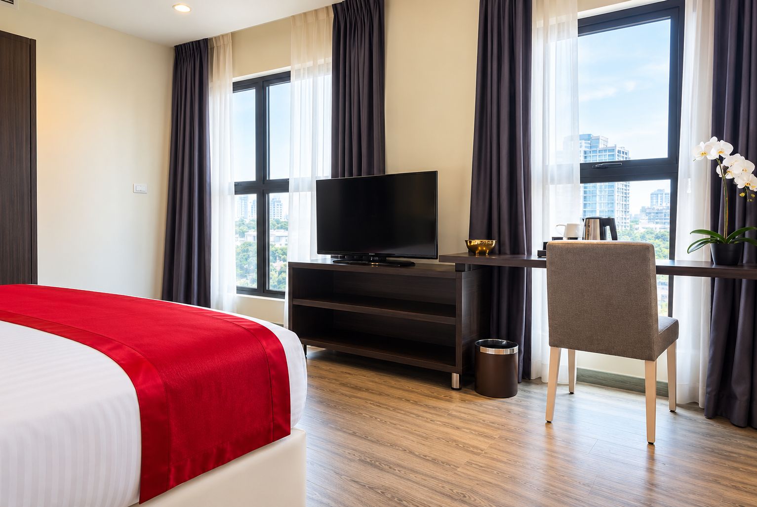 Habitación de hotel con una cama, un escritorio con televisor, una silla, una planta en maceta y vistas a la ciudad a través de amplias ventanas.