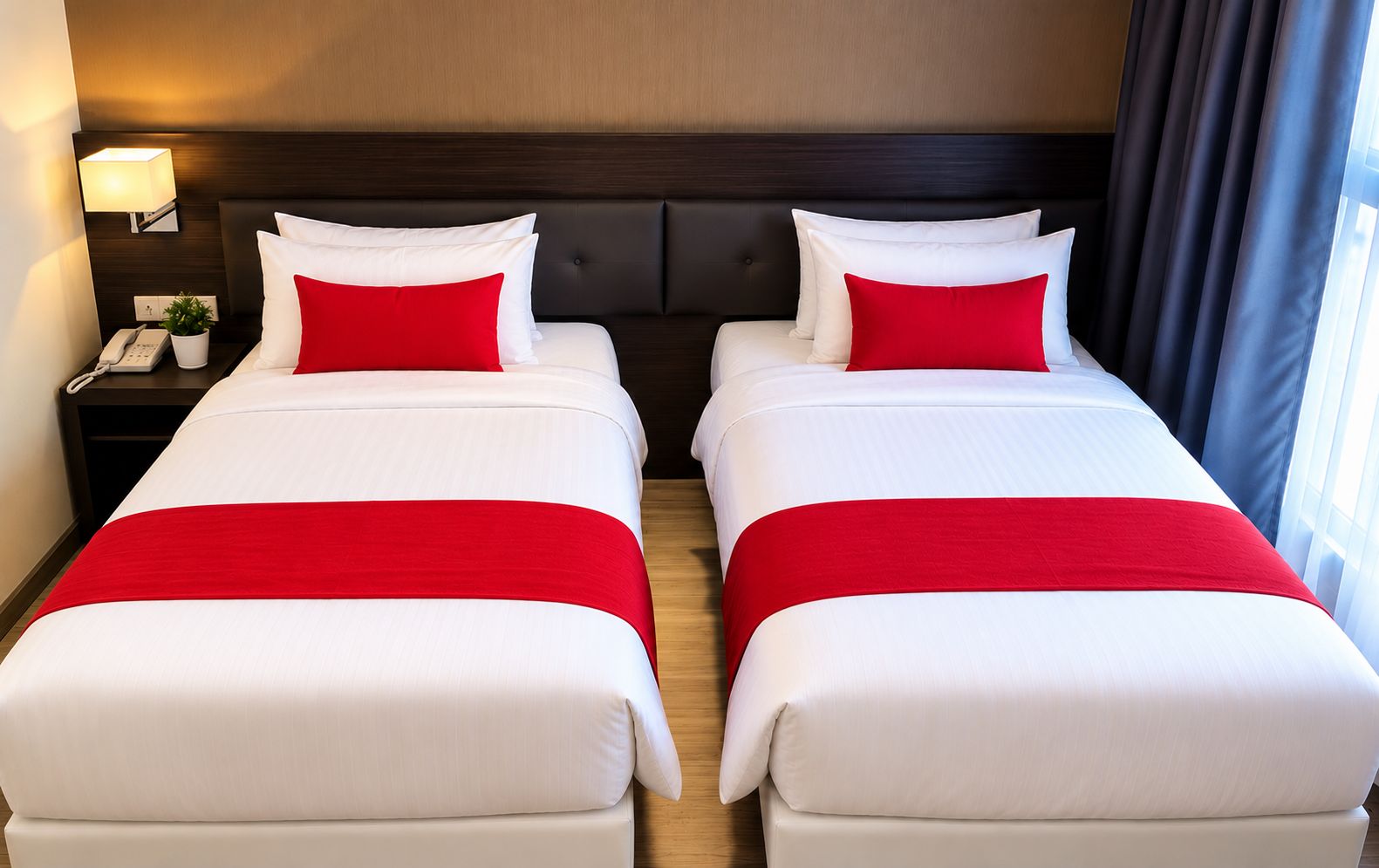 A hotel room with two beds, each with white bedding and red accents, a bedside table with a lamp, and a phone.