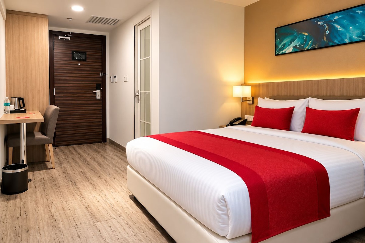 Een hotelkamer met een bed, een bureau, een stoel, een prullenbak, een deur en een schilderij dat aan de muur hangt.