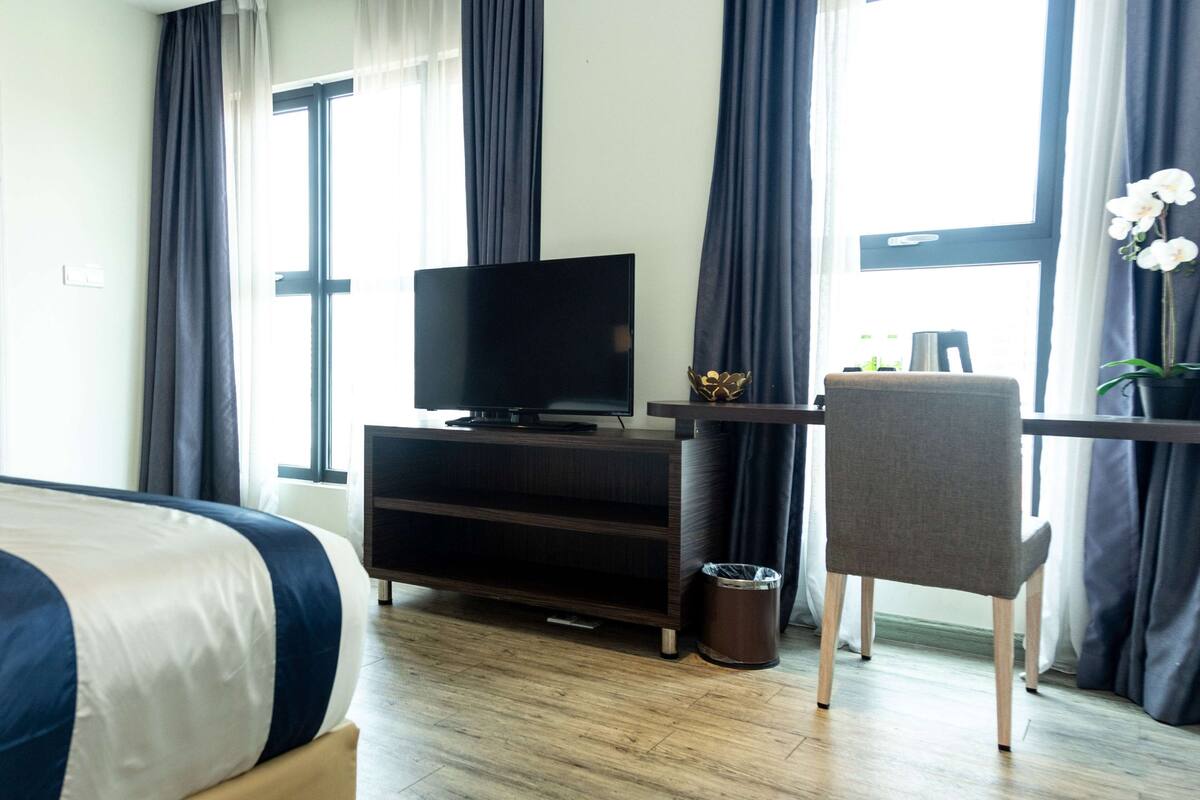 A hotel room with a bed, a TV on a wooden cabinet, a desk with a chair, and a window with curtains.