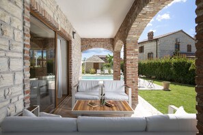 Un patio con un sofá blanco, una mesa de madera y un ventanal de vidrio que da a una zona de piscina.