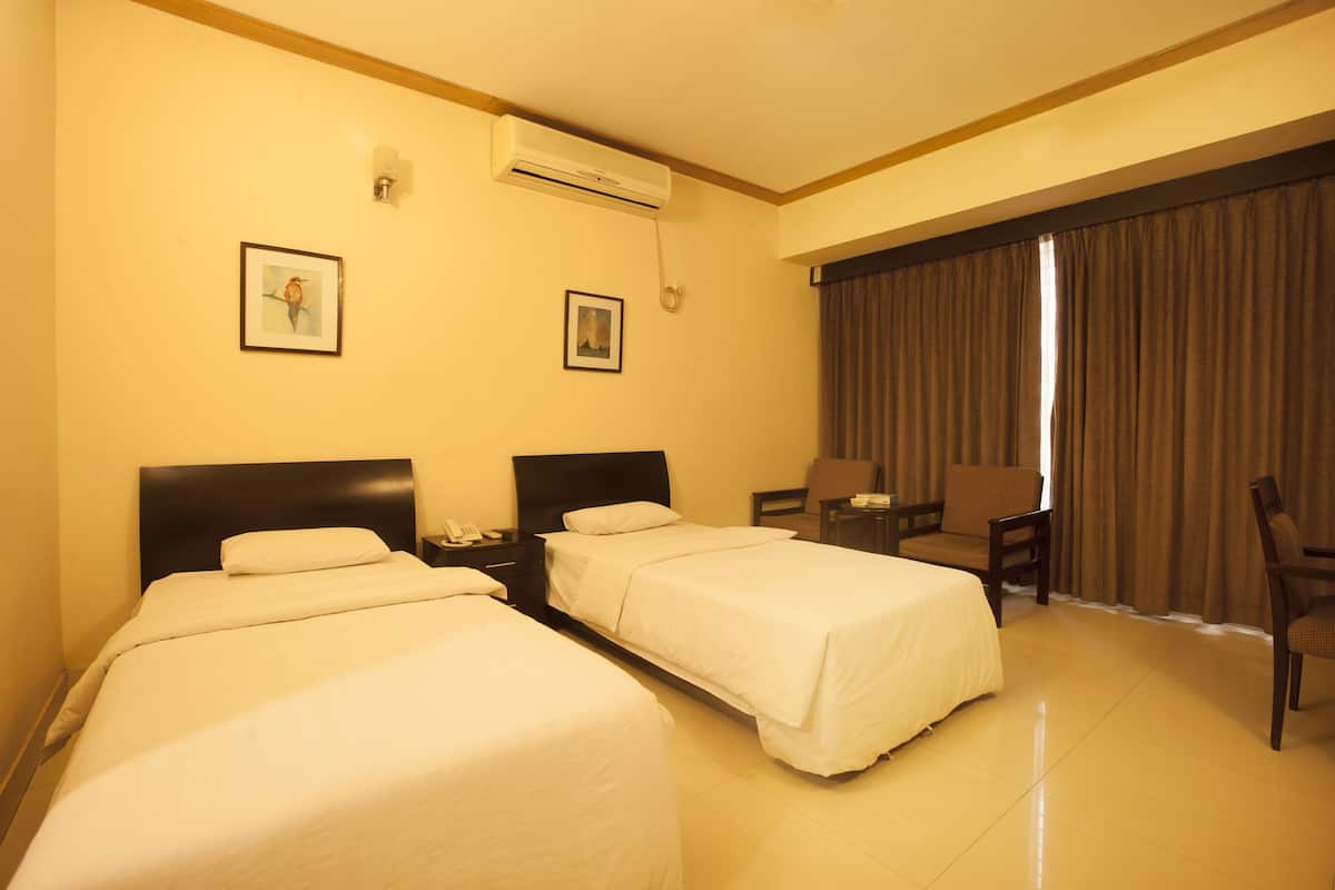 A hotel room with two single beds, a desk, a chair, a small table, an air conditioning unit, and two framed pictures on the wall.