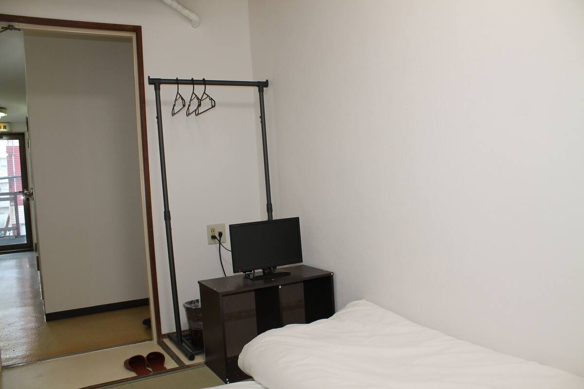 A bedroom with a bed, a desk with a computer, a clothes drying rack, and a door leading to another room.