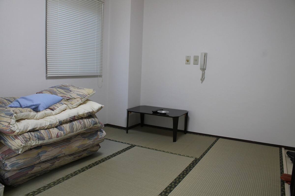A room with a bed, a small table, a wall socket, and a window with blinds.