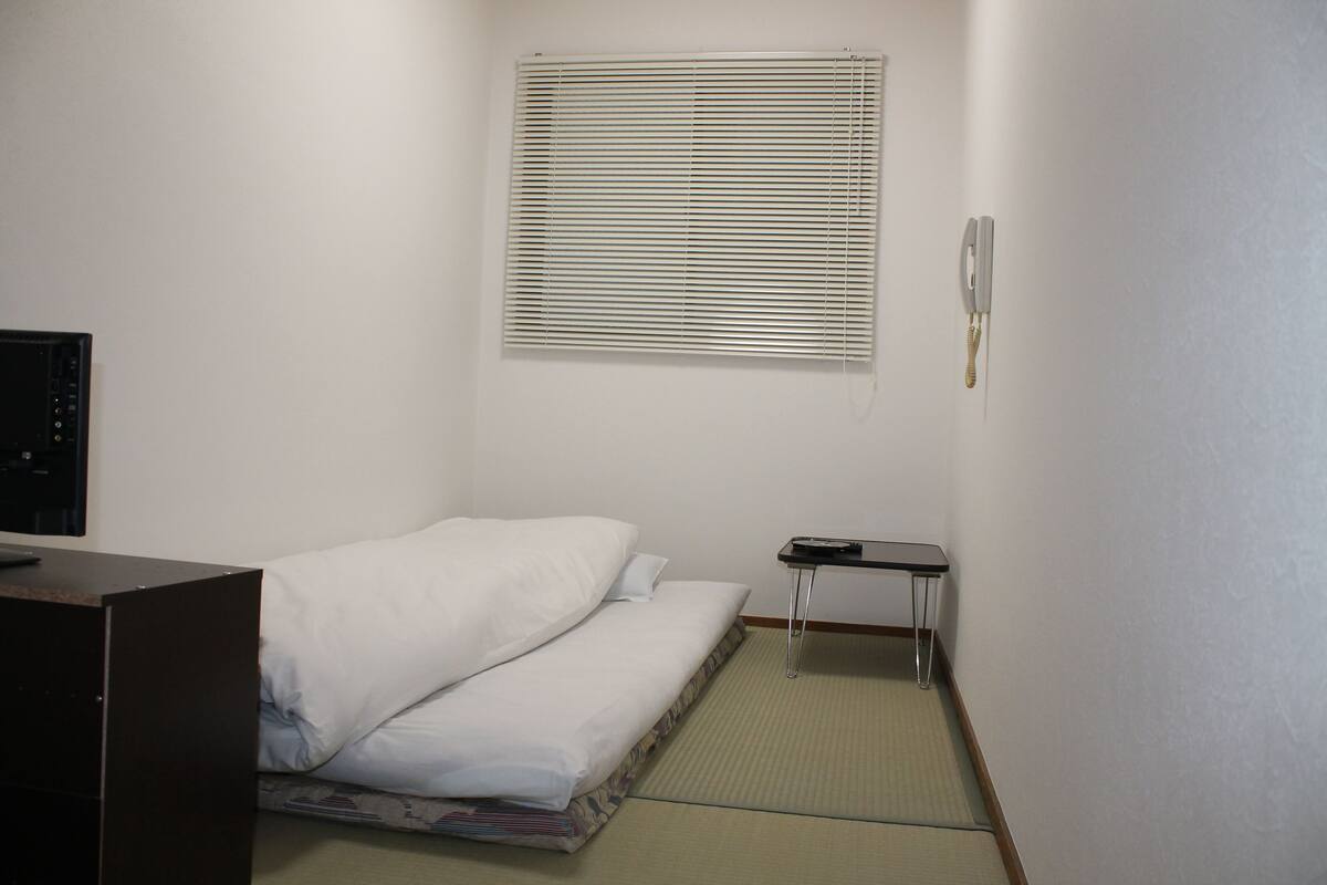 A small hotel room with a bed, a desk, a chair, a telephone, and a window with blinds.