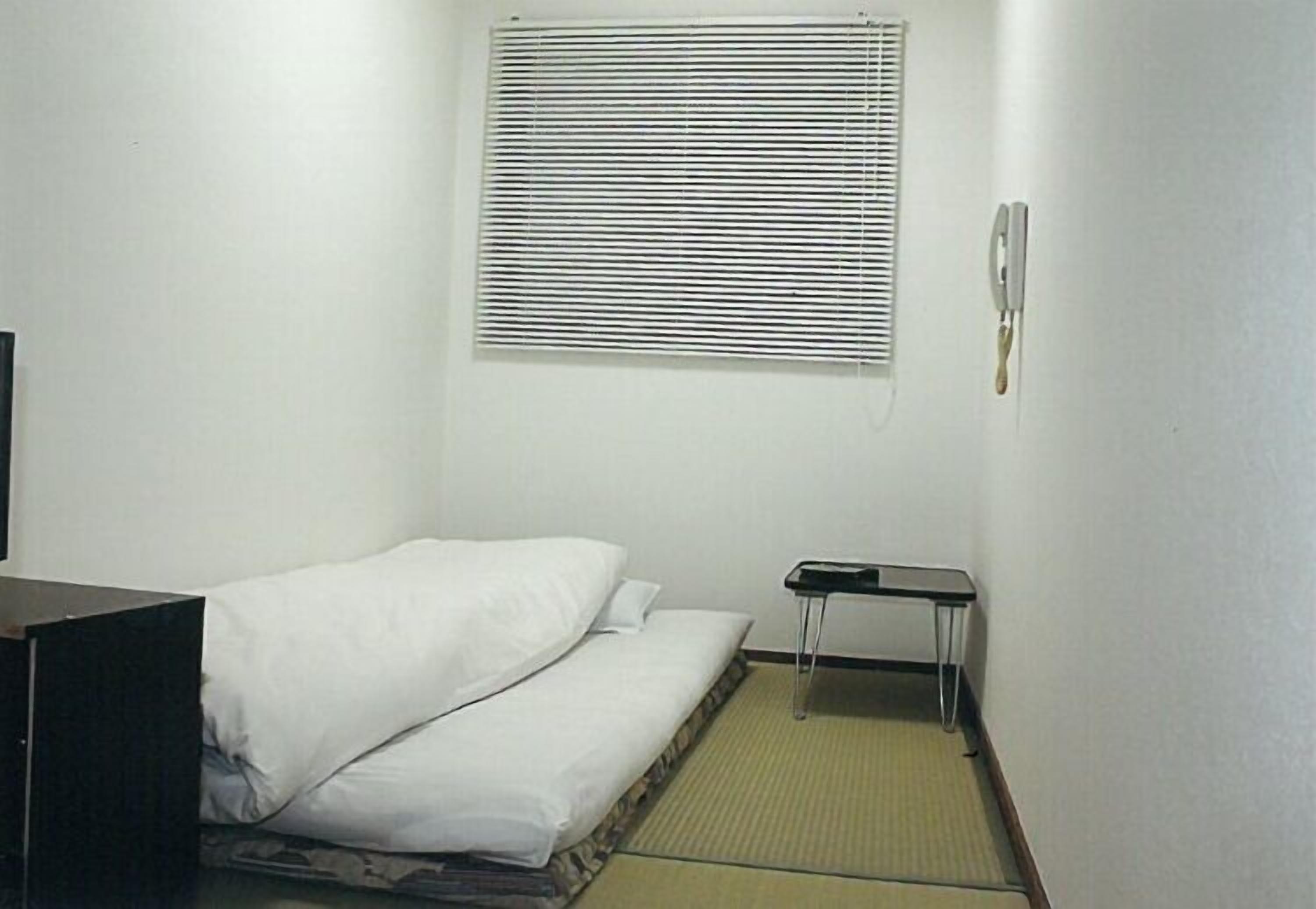A small, single-bed room with a window, a bedside table, and a wall-mounted phone.