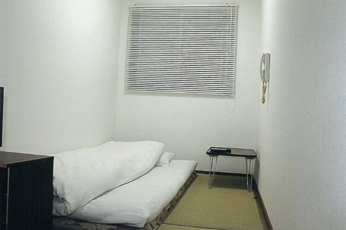 A small, single-bed room with a window, a bedside table, and a wall-mounted phone.