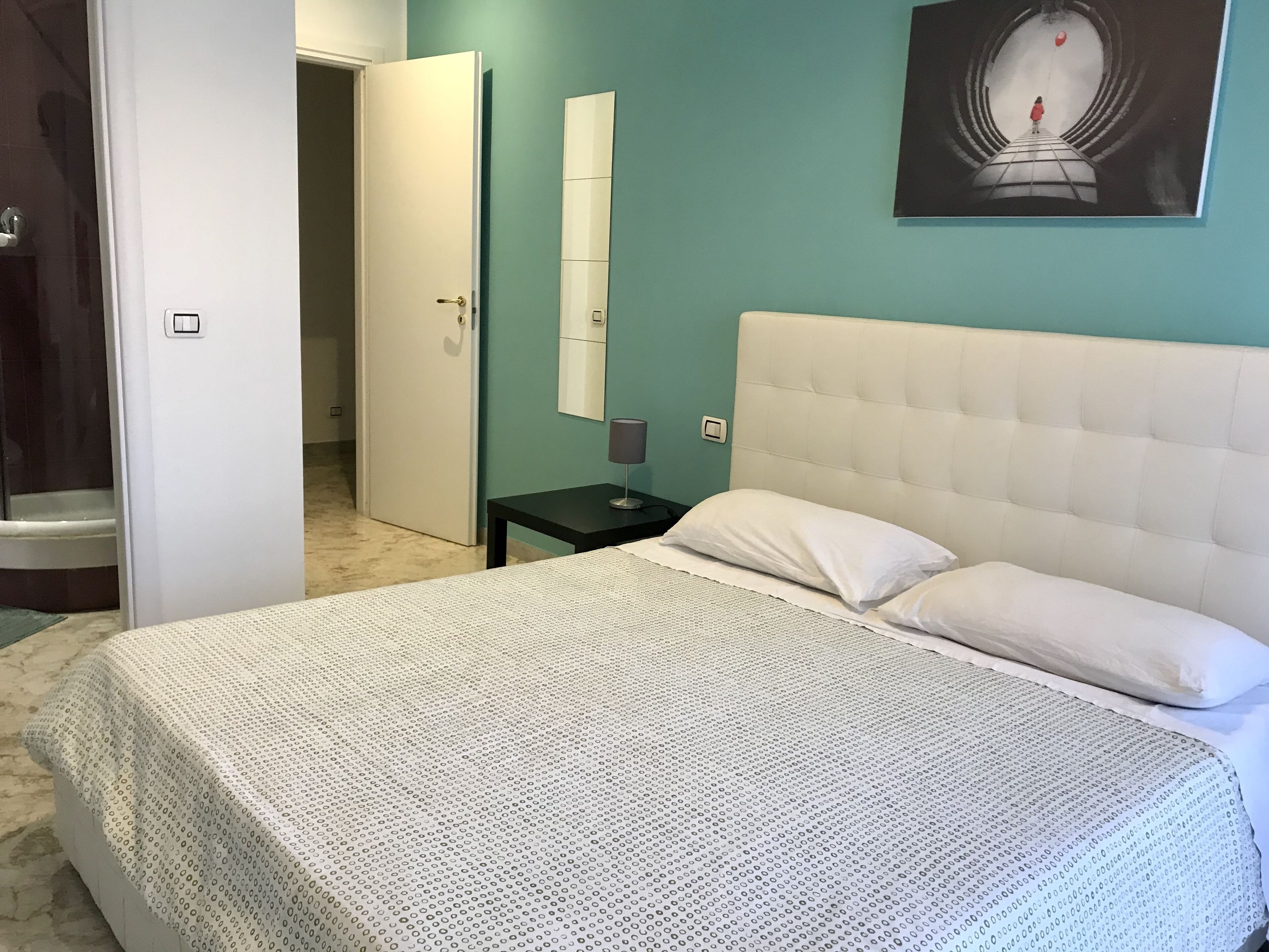 A bedroom with a white bed, a nightstand, a wall-mounted mirror, and a door leading to another room.