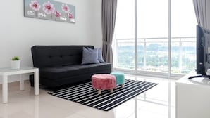A modern living room with a black sofa, a small white table, a potted plant, and a large window offering a city view. - Duet HomeStay (Seri Kembangan)