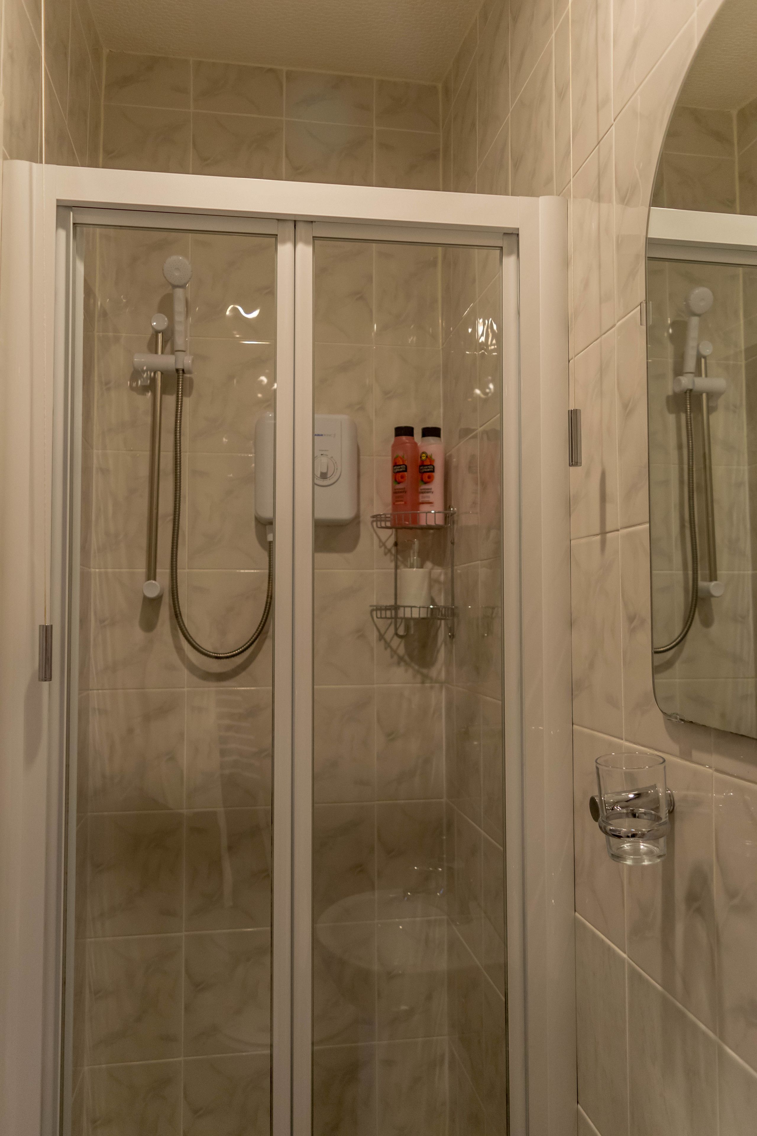 A glass-enclosed shower with a handheld showerhead, a wall-mounted control panel, and a shelf with toiletries.