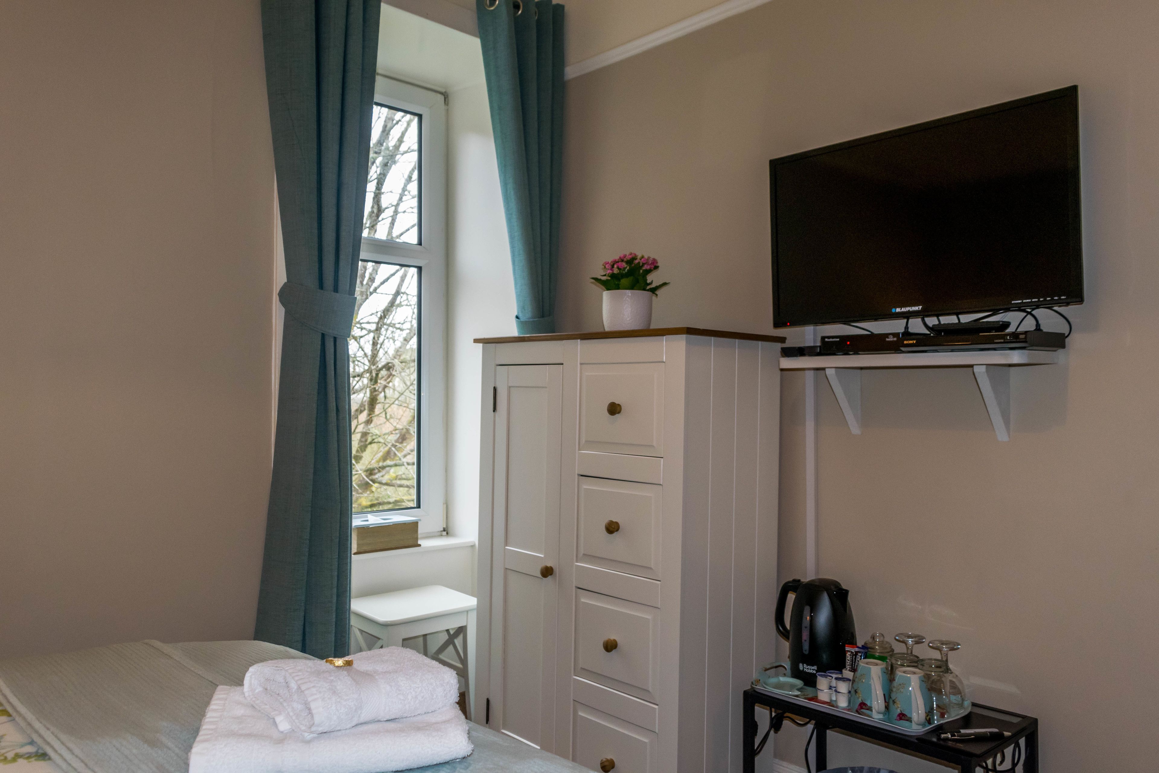 A hotel room with a bed, a TV mounted on the wall, a cabinet with drawers, a small table with a kettle and glasses, and a window with curtains.