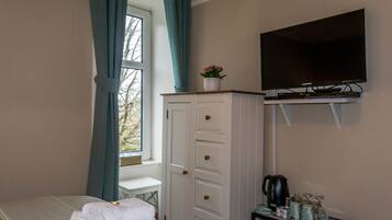 A hotel room with a bed, a TV mounted on the wall, a cabinet with drawers, a small table with a kettle and glasses, and a window with curtains.