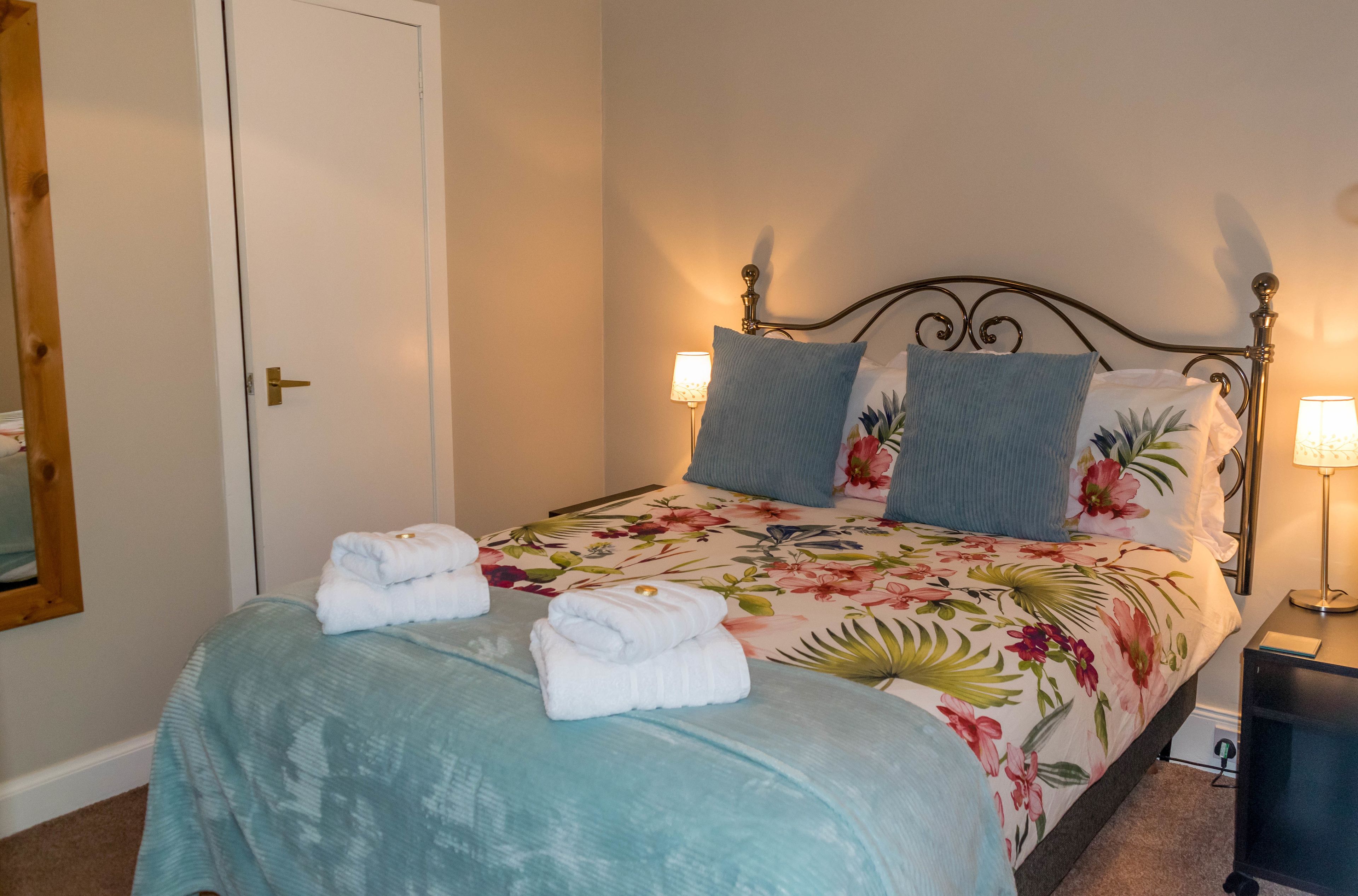 Una cama bien hecha con ropa de cama floral, dos almohadas y toallas dobladas sobre un cabecero.