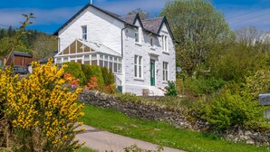 A white two-story house with a stone wall, a green door, and a small porch with a bench. - Tregortha (Tighnabruaich)