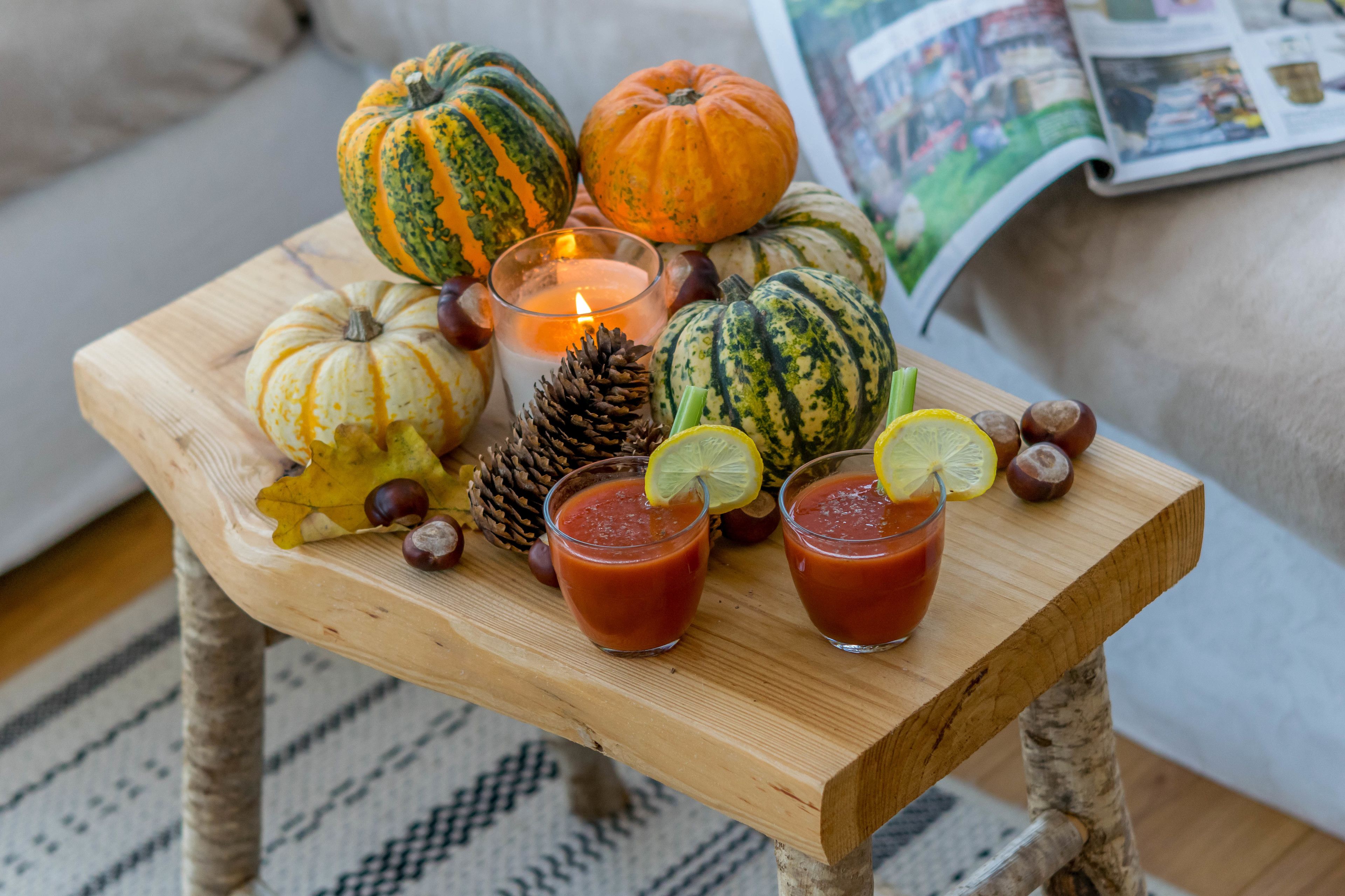 Una mesa de madera con varias calabazas, una vela, piñas y dos vasos de un brebaje rojo adornado con lima.